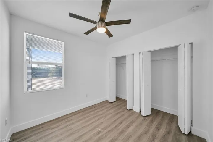 Wood-finish flooring, white walls, two bi-fold door closets, a window with blinds, and a ceiling fan with integrated lighting