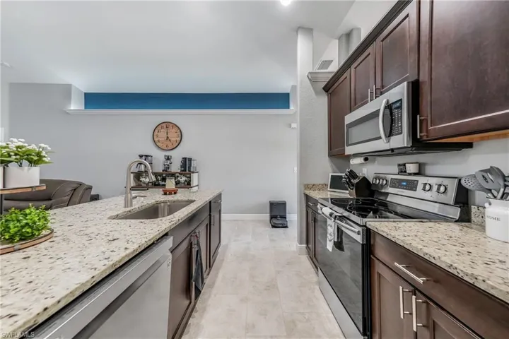 Kitchen with appliances with stainless steel finishes, a sink, dark brown cabinets, light stone countertops, and baseboards