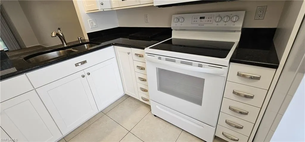 Kitchen with white electric range oven, dark stone counters, light tile patterned floors, and white cabinetry