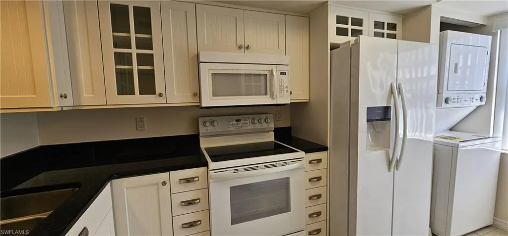 Kitchen featuring white appliances, glass insert cabinets, stacked washing machine and dryer, white cabinetry, and dark stone counters
