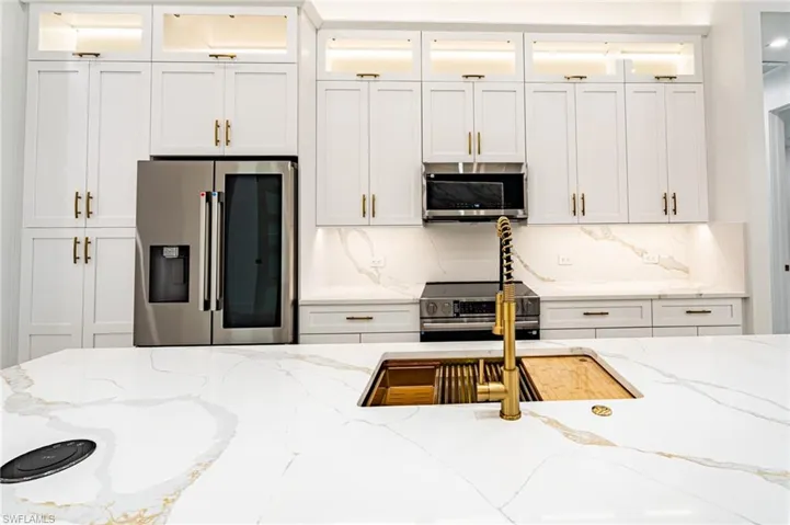 White shaker cabinetry with gold-tone hardware, stainless steel appliances, white countertop with gold veining, built-in sink with gold-tone faucet, and under-cabinet lighting