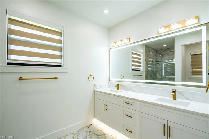 Dual vanity with white cabinetry and integrated sinks, under-cabinet lighting, large illuminated mirror, and gold-finish fixtures