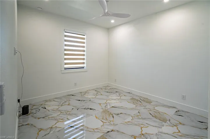 Bright, newly finished bedroom featuring polished stone-effect flooring with warm veining throughout, recessed ceiling lighting, ceiling fan, and a large transom window with motorized shades providing natural illumination and light control