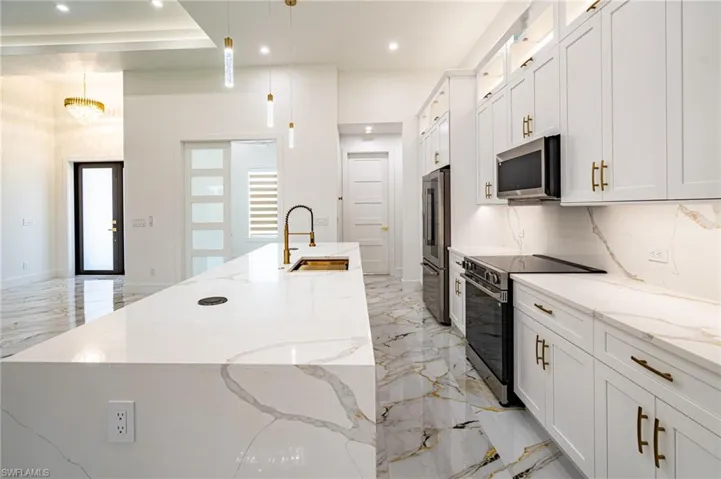 Spacious kitchen featuring an expansive island, white cabinetry with gold-toned hardware, undermount sink with a gooseneck faucet, integrated stainless steel appliances, and large-format porcelain tile flooring