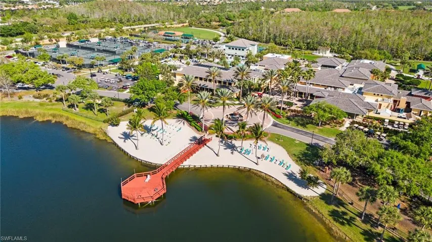 Lakefront property featuring a red dock, sandy beach area with lounge chairs, multiple buildings with tile roofs, palm trees, and tennis courts