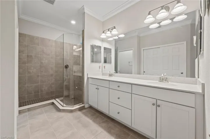 Spacious bathroom featuring a glass-enclosed shower with tile surround, a double vanity with ample storage, a large mirror, and crown molding