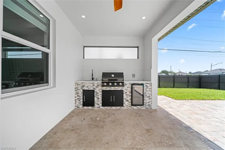 Covered outdoor kitchen featuring a built-in grill, integrated sink, beverage refrigerator, and tile backsplash