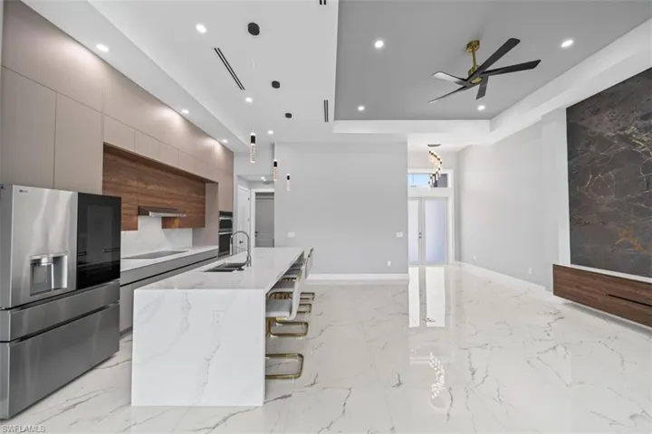 Open-concept living space with large format marble-look tile flooring, a modern kitchen island with waterfall edge, integrated stainless steel appliances, a recessed ceiling with accent lighting, and a feature wall with stone-look paneling