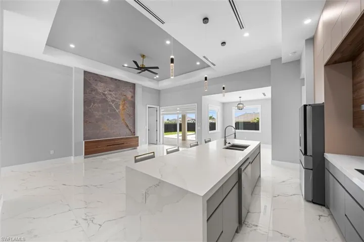 Spacious open-concept interior featuring large format marble-look tile flooring, a kitchen island with integrated sink, modern pendant lighting, a recessed ceiling with integrated lighting, and a feature wall with a wood-finish floating cabinet