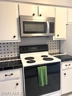 Kitchen featuring white electric range oven, stainless steel microwave, decorative backsplash, and white cabinets