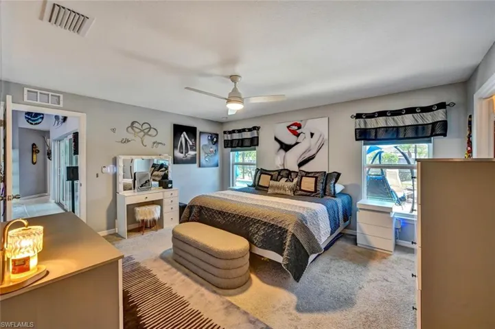Carpeted bedroom featuring a ceiling fan, two windows with valances, a built-in vanity with mirror, and a doorway opening to an interior hallway with tile flooring