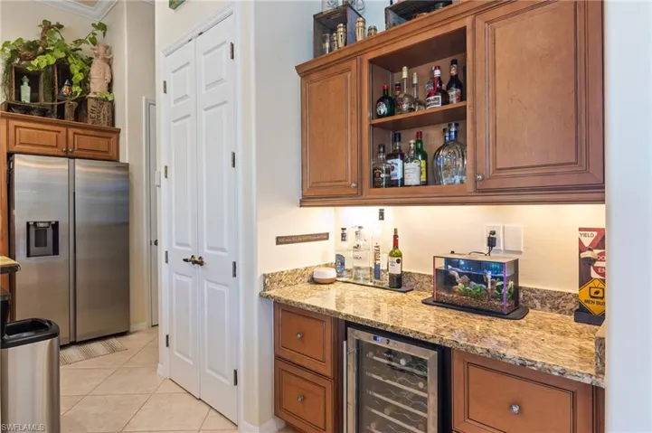 Bar with wine cooler, crown molding, stainless steel refrigerator with ice dispenser, light stone counters, and light tile floors