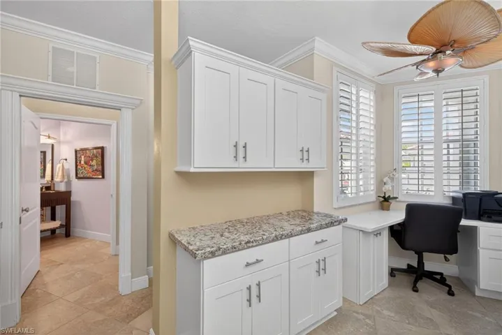This versatile space features built-in white cabinetry with a granite countertop, a dedicated desk area with ample natural light from windows with plantation shutters, and a ceiling fan