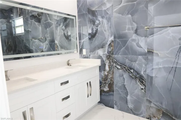 Full bath featuring double vanity, a marble finish shower, light marble finish floors, tile walls, and stone wall