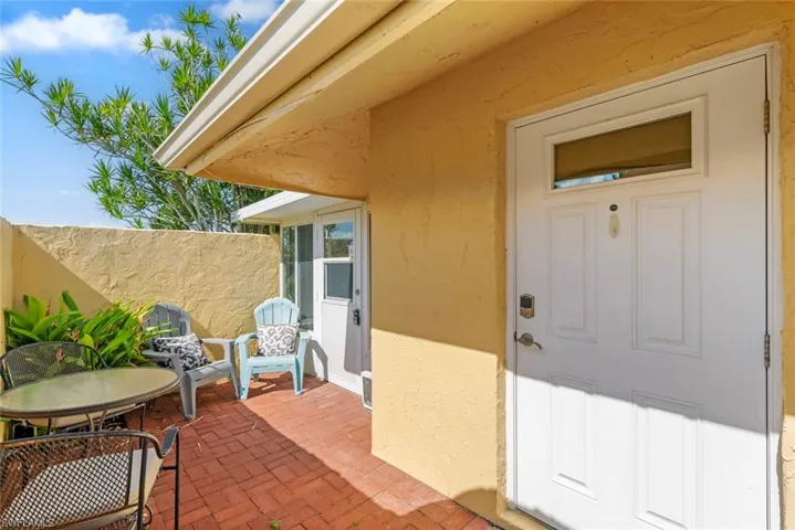 Inviting entry patio featuring a white paneled door with transom window, textured stucco exterior, red brick paving, and a garden bed with lush foliage