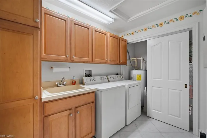 Clothes washing area with water heater, sink, light tile patterned floors, washing machine and dryer, and cabinets