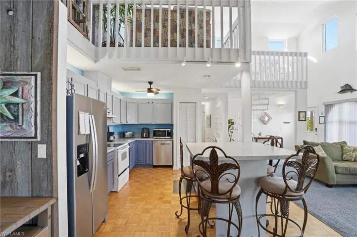 Kitchen with open floor plan, stainless steel appliances, a towering ceiling, a kitchen breakfast bar, and light countertops - Virtually Edited Image