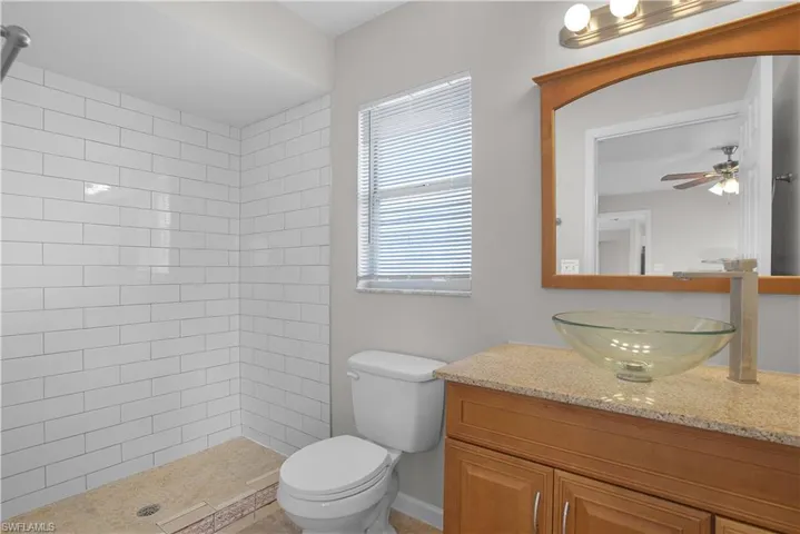 Bathroom featuring a walk-in shower with subway tile surround, a vessel sink, a wood-finish vanity with a light-toned countertop, and a window with horizontal blinds