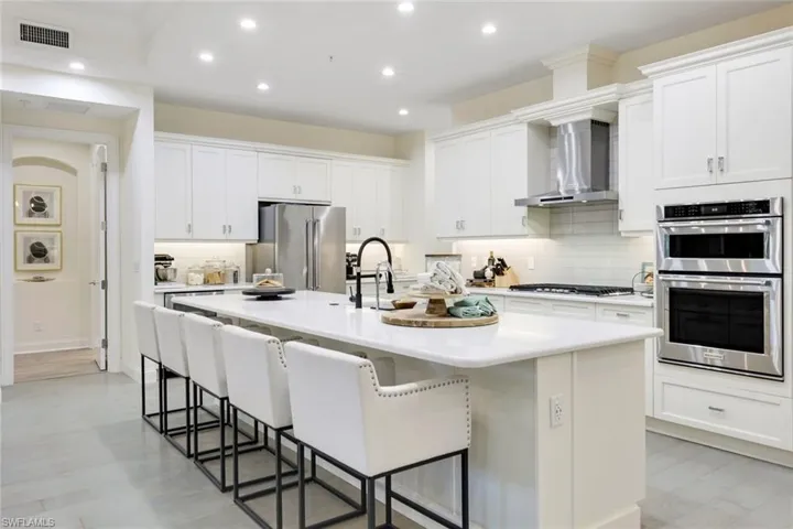Kitchen with backsplash, light hardwood / wood-style flooring, stainless steel appliances, wall chimney range hood, and an island with sink