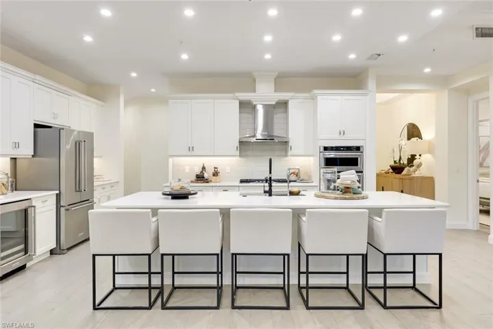 Kitchen with light wood-type flooring, tasteful backsplash, wall chimney exhaust hood, a center island with sink, and white cabinets
