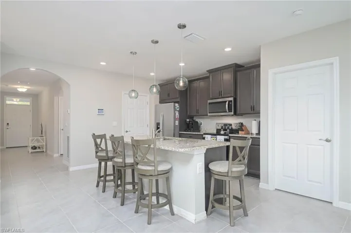 Kitchen featuring light stone countertops, pendant lighting, stainless steel appliances, dark brown cabinets, and a kitchen island with sink