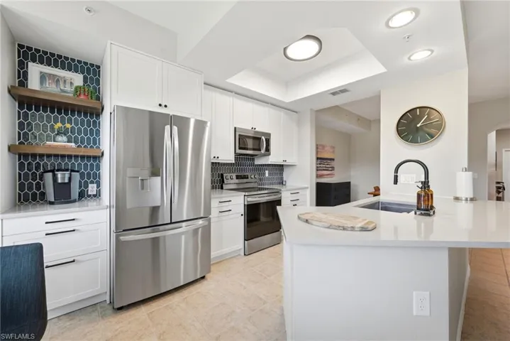 Kitchen featuring white shaker cabinetry, stainless steel appliances, quartz countertops, a dual-tone hexagonal tile backsplash, and wood-finish floating shelving