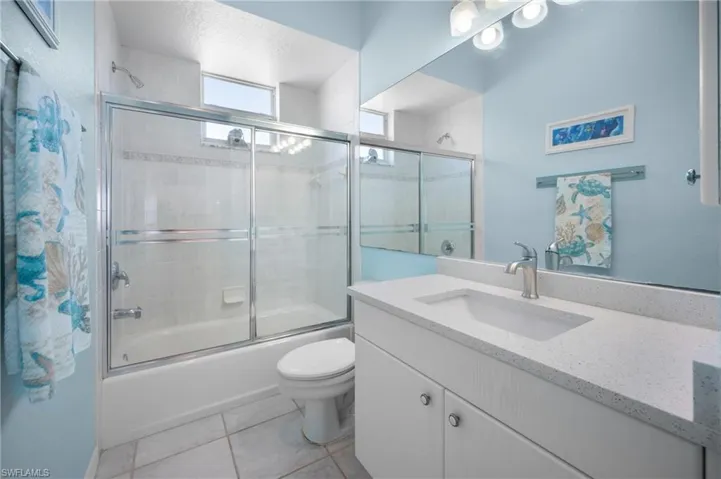 White vanity with an integrated sink and light-toned countertop, large wall-mounted mirror, and a bathtub with a sliding glass enclosure and tiled surround