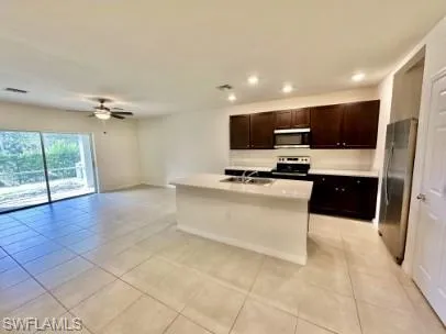 Open-concept kitchen and living area featuring tile flooring, a kitchen island with integrated sink, dark wood-finish cabinetry, stainless steel appliances, and a ceiling fan