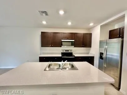 Spacious kitchen featuring an island with an integrated double basin sink, dark wood-finish cabinetry, light-toned countertops, and stainless steel appliances including a refrigerator, range, and microwave