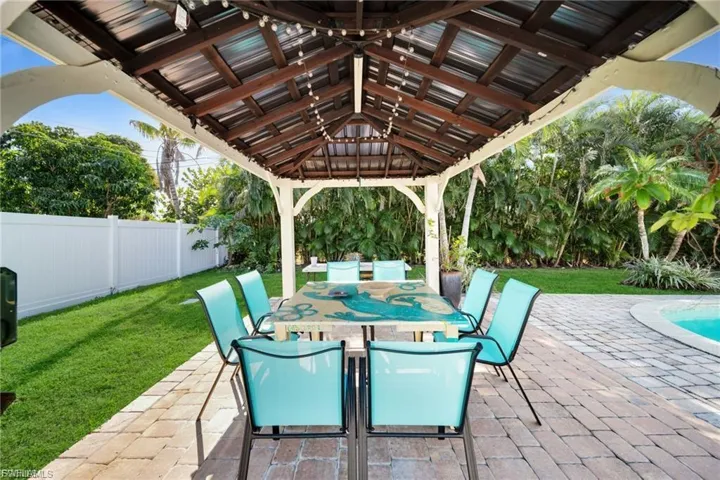 Expansive covered patio featuring a wood-beam pergola with string lighting, paver flooring, and abundant tropical landscaping