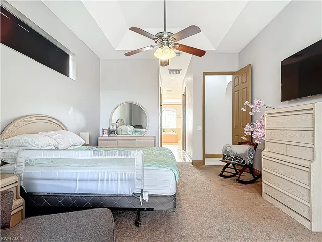 Carpeted bedroom with ensuite bathroom, ceiling fan, and a tray ceiling