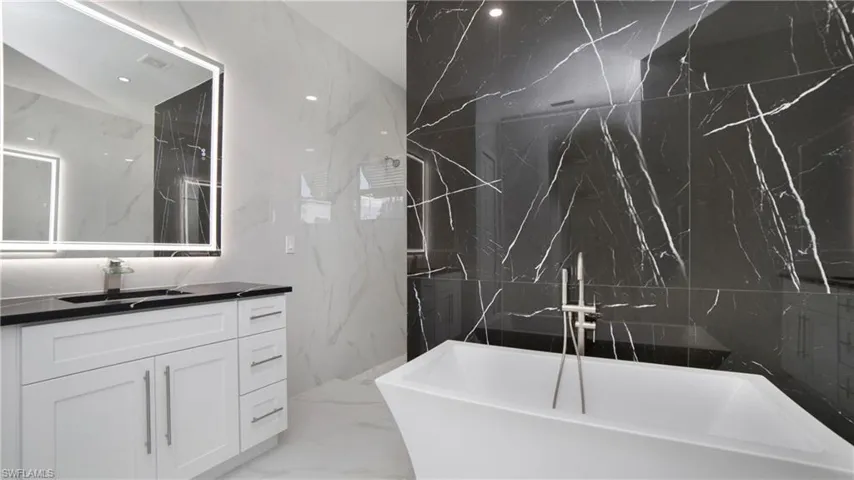 Modern bathroom featuring a standalone soaking tub with floor-mounted faucet, black and white veined tile walls, a white vanity with shaker-style cabinetry, a dark countertop, and an illuminated mirror