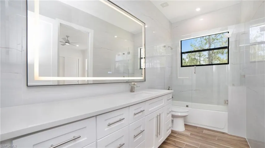 Contemporary bathroom featuring a spacious vanity with white cabinetry and a light-toned countertop, an illuminated rectangular mirror, a white bathtub with a glass shower panel, and wood-finish tile flooring