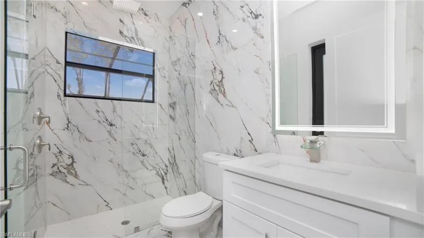 Bathroom featuring full-height marble-style tiled walls, a glass-enclosed shower with a rainfall showerhead, a single vanity with a white countertop and base cabinetry, and an illuminated mirror