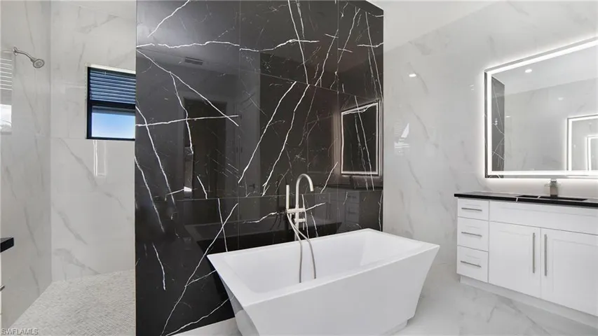 Contemporary bathroom featuring a freestanding soaking tub, a black tiled accent wall, and a custom vanity with an illuminated mirror