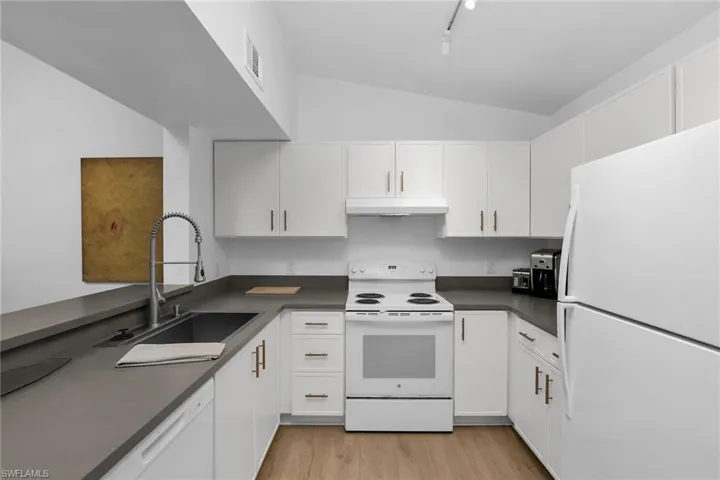 Kitchen featuring white appliances, under cabinet range hood, lofted ceiling, a sink, and dark countertops