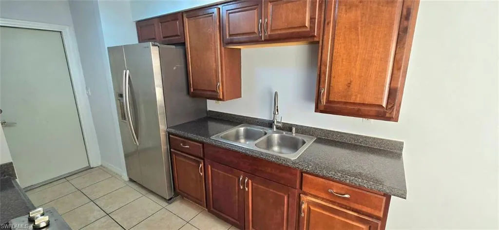 Kitchen featuring dark countertops, light tile patterned floors, stainless steel refrigerator with ice dispenser, and bold wood finish cabinetry