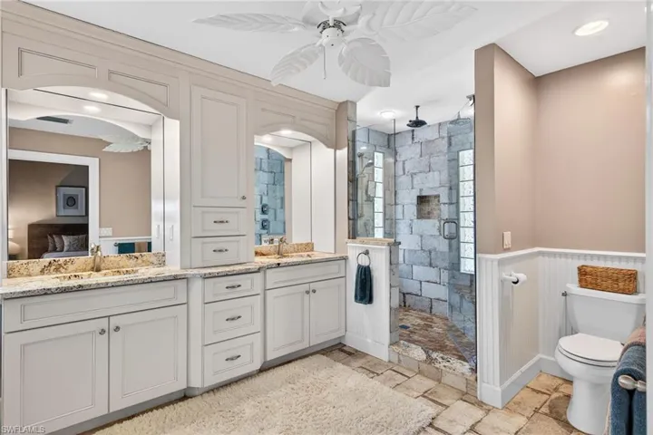 Full bathroom featuring double vanity, a stall shower, stone tile floors, ceiling fan, and wainscoting