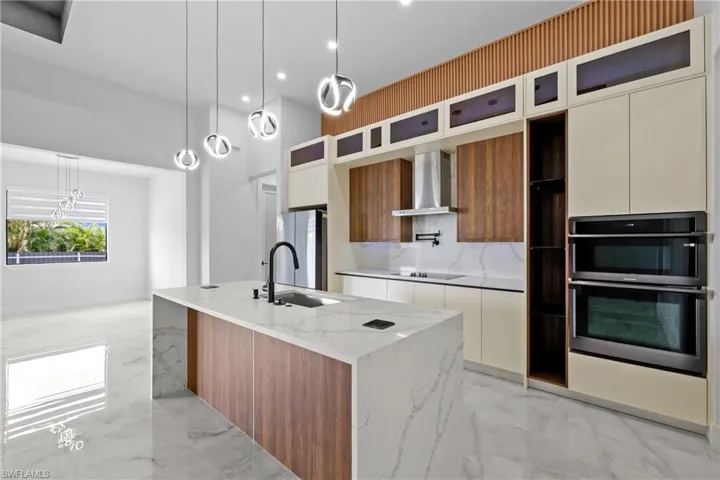 Kitchen featuring wall chimney range hood, white cabinetry, stainless steel appliances, pendant lighting, and a center island with sink