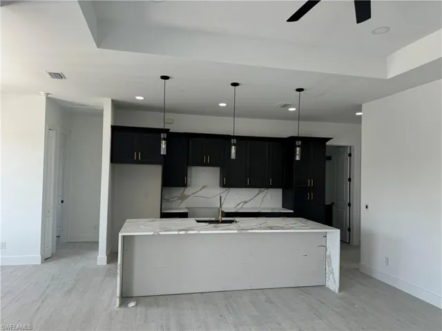 Kitchen with tasteful backsplash, ceiling fan, an island with sink, light stone countertops, and hanging light fixtures