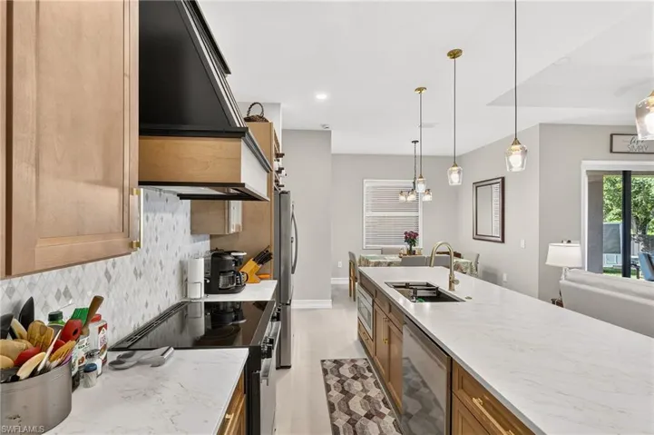 Kitchen featuring light wood-finish cabinetry, a diamond-patterned tile backsplash, a built-in range hood, a kitchen island with an undermount sink, and pendant lighting