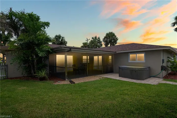 Back of property at dusk featuring a hot tub, stucco siding, a lawn, and roof with shingles