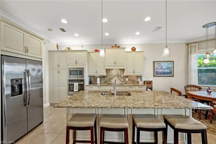 Expansive kitchen featuring a large island with an undermount sink and pendant lighting, light-toned cabinetry, stainless steel appliances, a built-in oven, and granite countertops with a matching backsplash