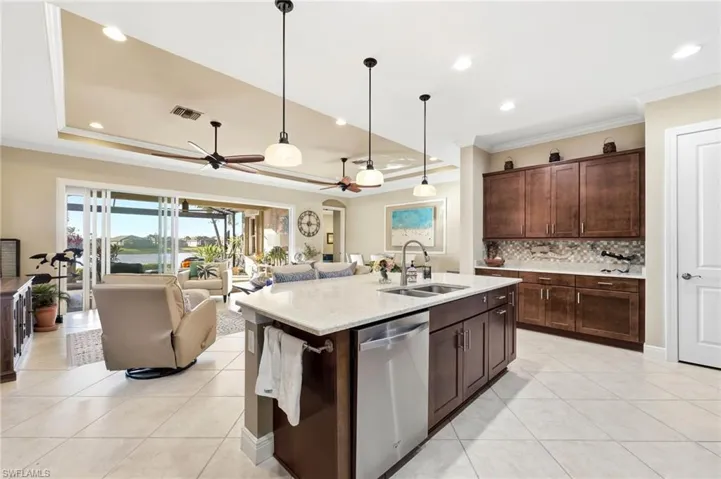 Kitchen featuring open floor plan, dark wood finish cabinets, light stone countertops, dishwasher, and a kitchen island with sink