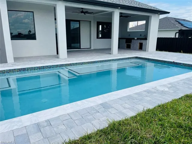 In-ground swimming pool with decorative tile inlay, covered patio featuring ceiling fans, and an outdoor kitchen area with stone-finish base