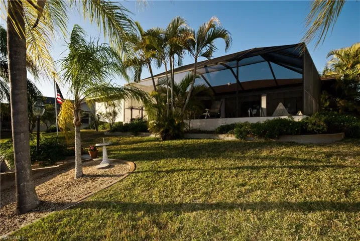 Exterior featuring a screened enclosure with glass panels, mature palm trees, landscaped garden beds, and a manicured lawn