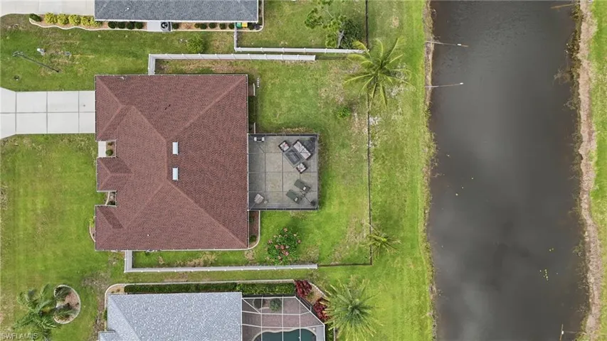Expansive aerial view showcasing the property's perimeter, a brown shingle roof, a concrete patio area, a manicured lawn, and a water feature bordering the property