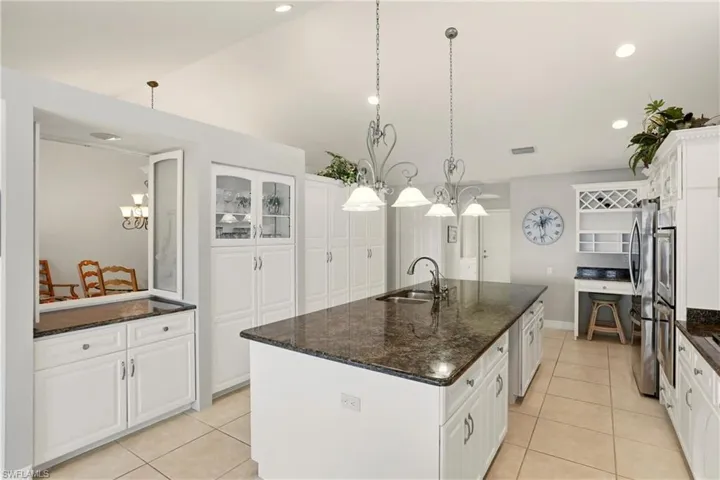 The kitchen features an oversized  island , an abundance of white cabinetry with granite countertops, hanging light fixtures, and vaulted ceiling.