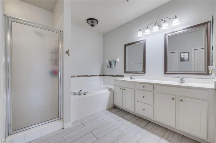Spacious bathroom featuring a dual vanity with white cabinetry, a built-in soaking tub, a separate shower with a frosted glass enclosure, and light wood-finish plank flooring