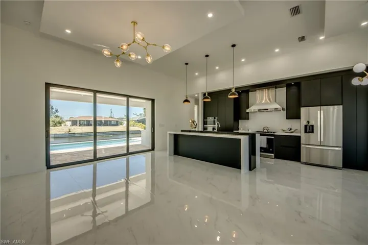 Kitchen with hanging light fixtures, wall chimney range hood, stainless steel fridge, an island with sink, and a chandelier